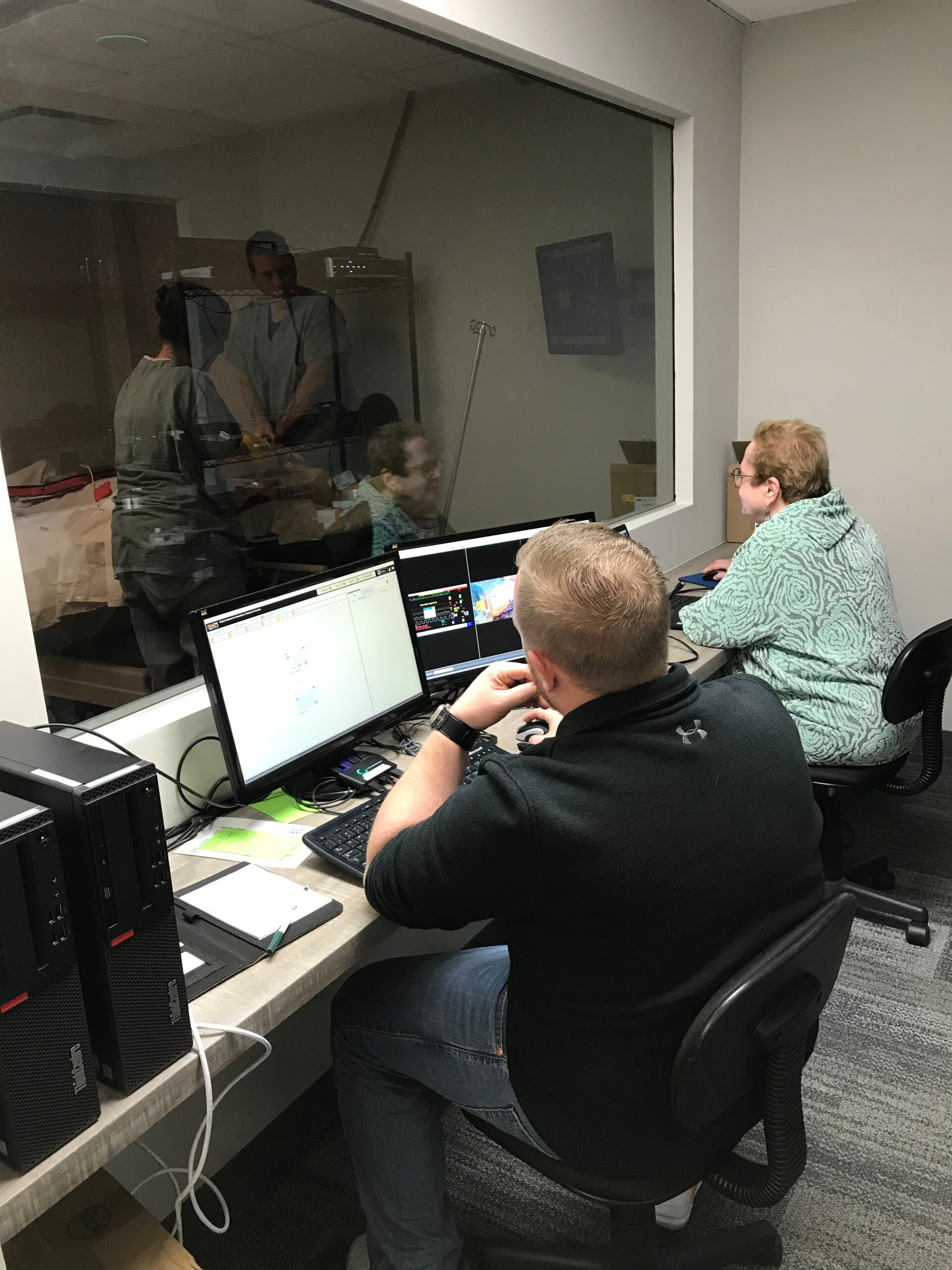 Two people work at a desk with multiple monitors in a control room; a glass window shows a person in scrubs inside a lab.