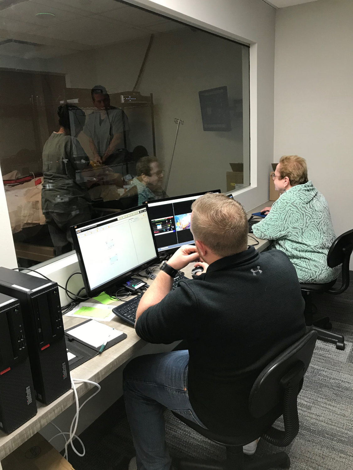 Two people sit at a desk with multiple computer monitors in an office, looking at screens; a glass window shows another room with a person standing by equipment.