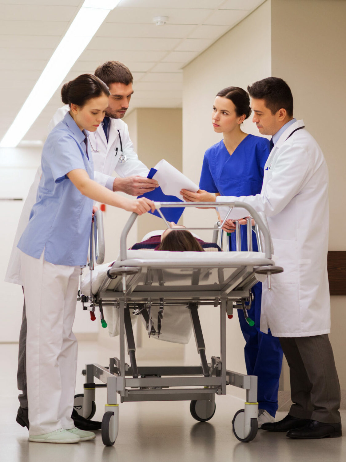 Medical team in scrubs and white coats gathered around a patient on a hospital stretcher, reviewing a chart in a hallway.