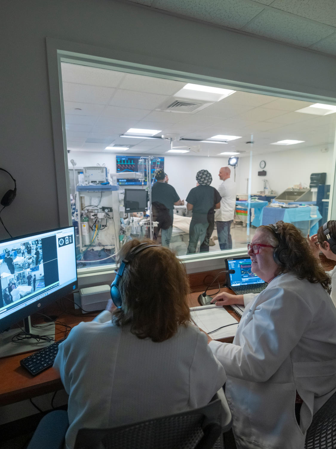 Doctors in a control room with headsets watching a surgical team through a glass window, monitors on desks show patient data.