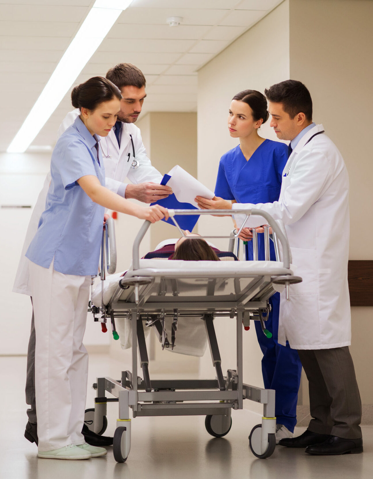 Medical team of four clinicians (two men in white coats, two women in scrubs) review documents beside a patient on a hospital stretcher in a bright corridor, coordinating care.