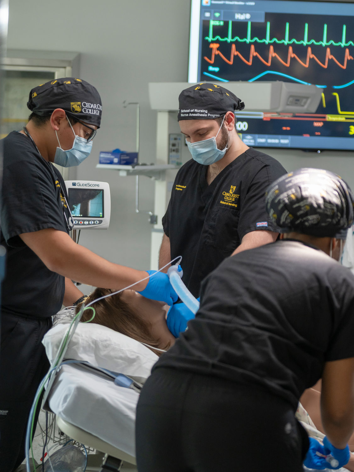 An anesthesiology team in scrubs and masks secures the patient’s airway with a breathing tube while a monitor shows vital signs in the background.