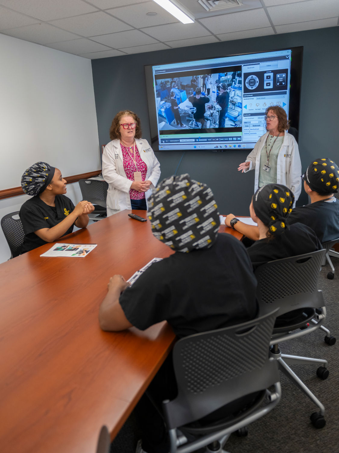 Two instructors in white coats lead a classroom discussion for students in black scrubs around a long wooden table; a large monitor displays medical images in the background.