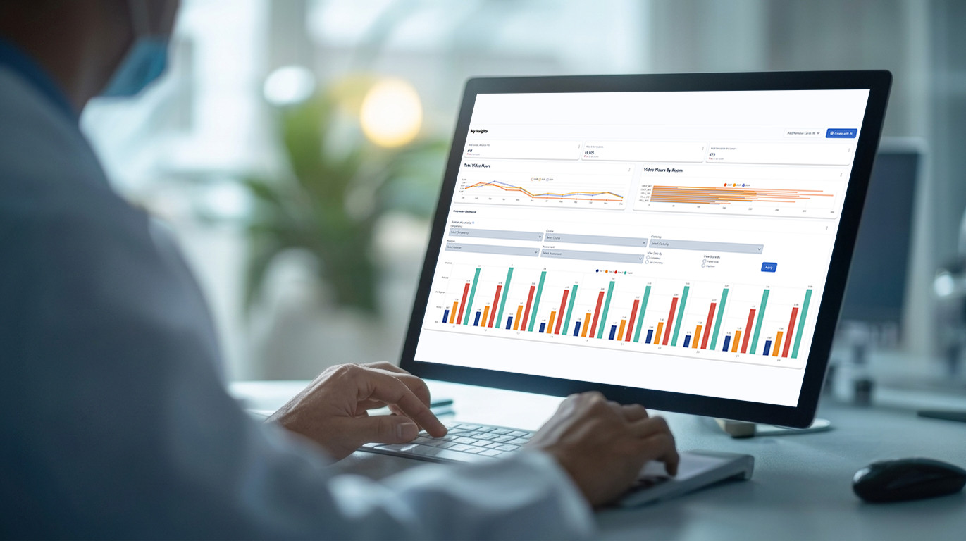 Person typing at a desk while a monitor shows a multi-panel data analytics dashboard with charts and graphs.