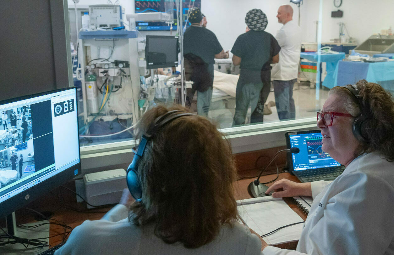 Two clinicians in a control room sit at desks with headsets and monitors, while an operating room with a surgical team is visible through a glass wall behind them.